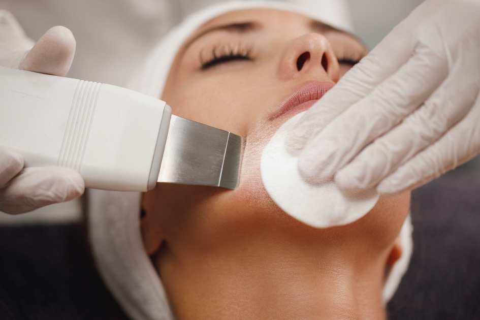 Centro Belleza Ponferrada Una persona está recibiendo un tratamiento facial. Están acostados con los ojos cerrados. Un profesional con guantes blancos sostiene una herramienta contra la mejilla de la persona mientras usa un algodón para limpiar su piel. La persona lleva una diadema y parece relajada. Estética