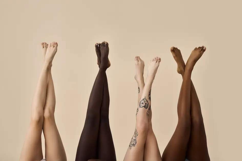 Centro Belleza Ponferrada Se muestran cuatro pares de piernas levantadas y cruzadas en los tobillos sobre un fondo beige neutro. Las piernas varían en tonos de piel y un par presenta varios tatuajes. La imagen se centra en la diversidad y el arte corporal. Estética