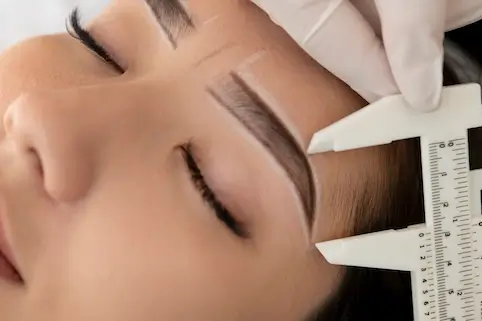 Centro Belleza Ponferrada Primer plano de una persona con los ojos cerrados mientras una mano enguantada usa hábilmente un calibrador para medir las cejas. La piel está marcada con líneas blancas, lo que indica la preparación para un tratamiento de cejas en el centro de belleza. Estética